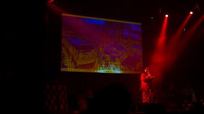 Performer standing on stage within a red spotlight, against a dark environment and a projector behind them.