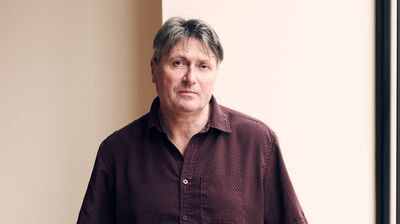 Simon Armitage standing in front of a wall in a red shirt.