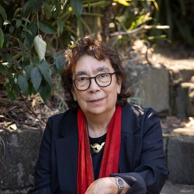 A woman with glasses and a red scarf sits on stone steps surrounded by greenery, smiling into camera.