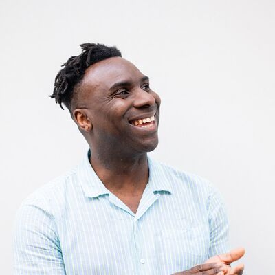 A man smiles and claps his hands together, wearing a light blue shirt against a plain background.