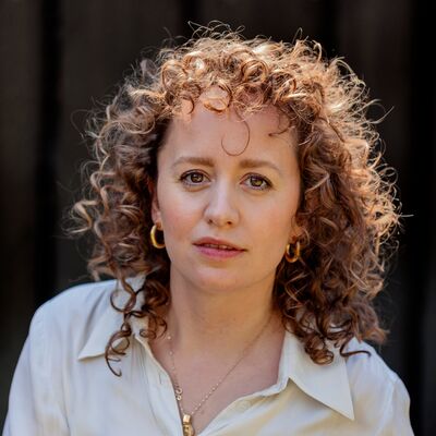 A woman with shoulder length curly hair wearing a white shirt against a black background.