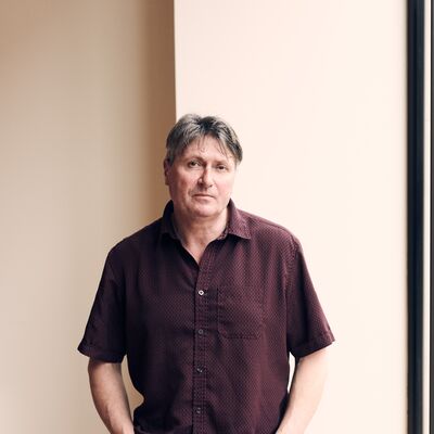 Simon Armitage standing in front of a wall in a red shirt.