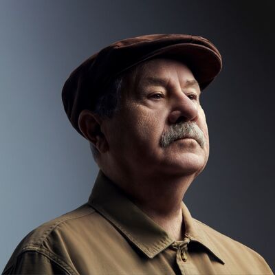 A man with a moustache wearing a brown flat cap and light brown shirt looking to the right