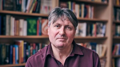 In front of a bookshelf, a man with a red pattered shirt stares into the camera with an mild expression