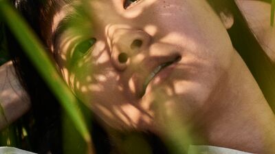 Someone lying down with grass and other foliage causing shadows on their face, wearing a white top