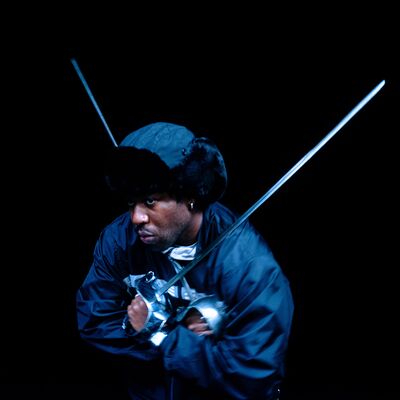 A Black man wearing a fur-lined aviator hat and a black jacket wields two fencing foils against a dark background.