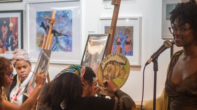 Two women painting at easels whilst another stands at a microphone, with framed art hanging in the background.