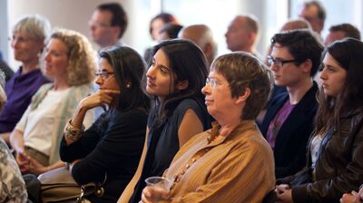 A audience is seated, attentively listening to a presentation with various expressions within a well lit setting