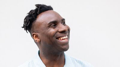 A man smiles and claps his hands together, wearing a light blue shirt against a plain background.