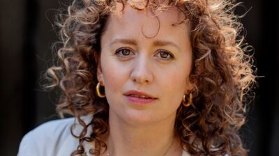 A woman with shoulder length curly hair wearing a white shirt against a black background.