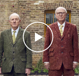 A still from a video featuring Gilbert, a shorter man with a green tweed suit, and George, a taller bespectacled man with a rusty tweed suit.