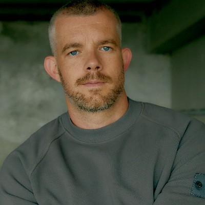 Someone with blue eyes and short grey brown hair wearing a grey jumper with their arms folded