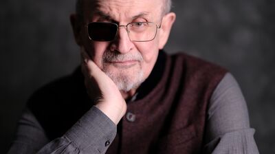 Salman Rushdie wearing a maroon waistcoat and grey shirt with his head in his hand.