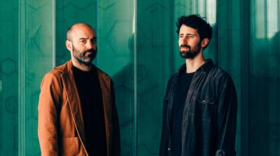Two people in front of a teal wall, on the left they have a shaved head and wear an orange jacket, on the right they have dark hair and a blue denim jacket