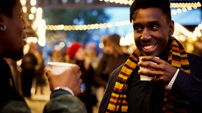 A man smiling at the Winter Market holding a cup of mulled wine, wearing a stripy orange scarf.