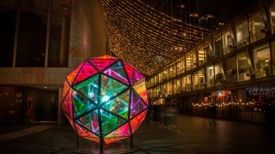 A vibrant, large, geometric sculpture made of multicolored glass panels glows at night under a canopy of string lights.
