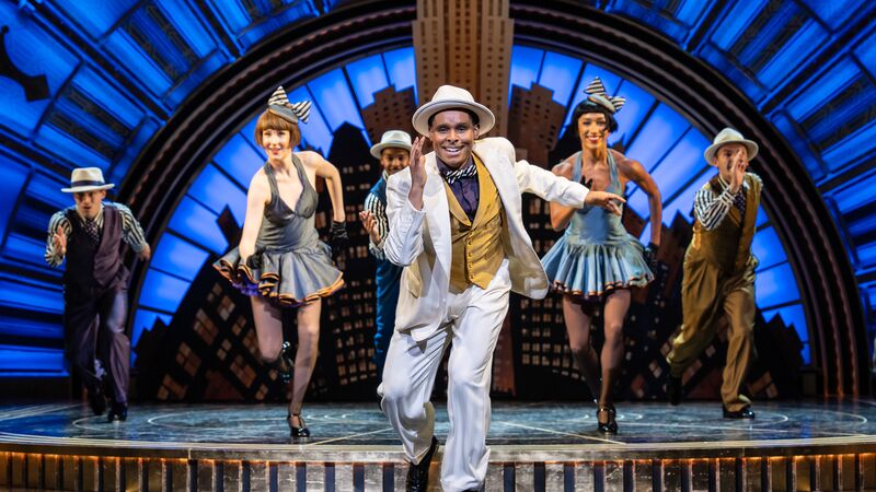 A theatrical performance with a male dancer in a white suit and hat leading a line of five dancers in 1920s-style costumes against a blue Art Deco backdrop.