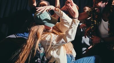 Dancers all moving together with one person in front wearing a white shirt and green cap with arms over her head