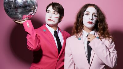 Two people dressed in pink suits with white mime makeup on their faces, one holding a disco ball