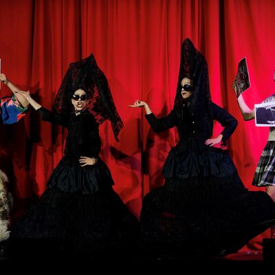Performers in outfits in front of a red curtain some holding news papers, two in black dresses and pointy hats