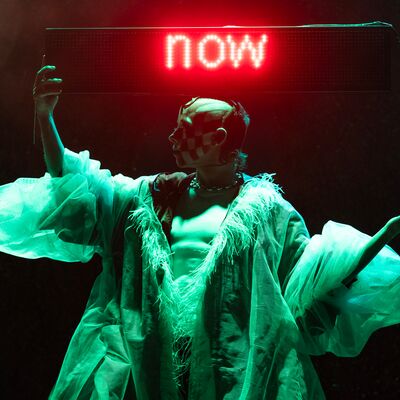 Performer wearing an extravagant outfit with puffy sleeves raising their arms and a ren LED sign above says 'NOW'