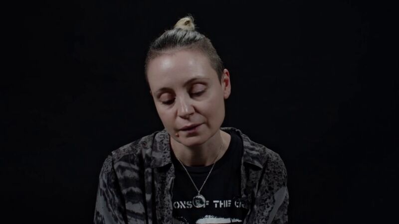Fran Lock, a woman with her blond hair pulled back and shaved at the sides, looks down whilst reading from their TS Eliot Prize shortlisted collection against a black background