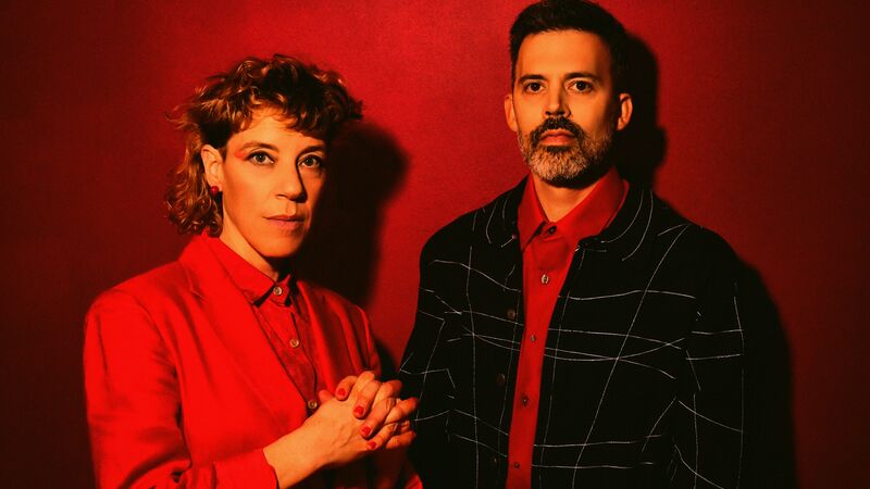 Two people one wearing a red suit and the other wearing a black suit against a red background