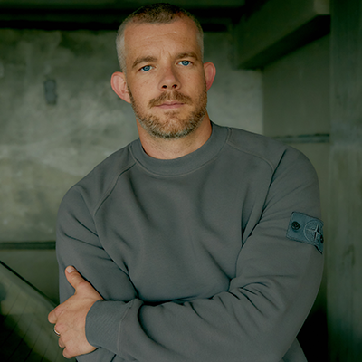 Someone with blue eyes and short grey brown hair wearing a grey jumper with their arms folded