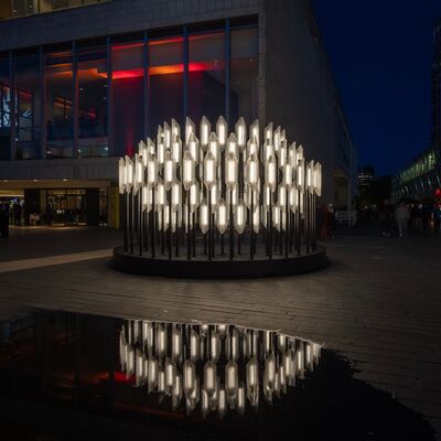A striking outdoor light installation of a ring of glowing, upright crystalline shapes reflected in a puddle on a dark city square.