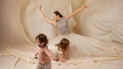 An image of the artist knelt down in a dress that billows out to cover the whole environment, with two young children running about the space.