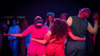 Three performers dance together, with their arms around each other's waists, drenched in pink light.