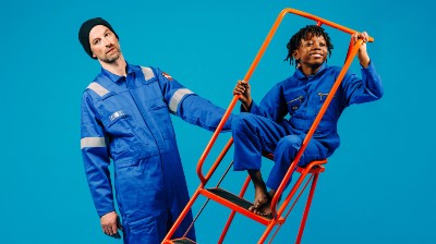 Two performers dressed in blue boiler suits stand in space together; a man holding an orange ladder which a small boy is sat in.