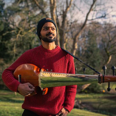 Soumik Datta standing outdoors holding his traditional instrument and wearing a red jumper.