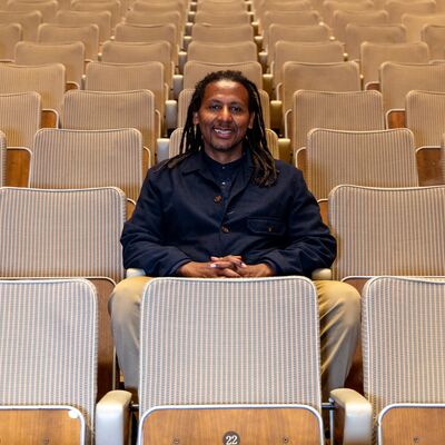 A Black man with long dark hair sits smiling among the seats of the Royal Festival Hall