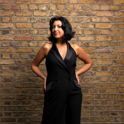 A woman wearing a black jumpsuit, stood against a brick wall