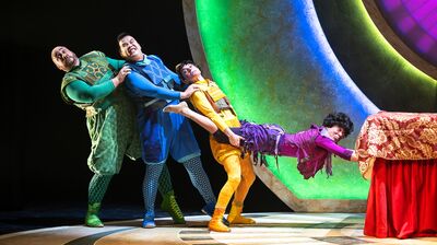 Four brightly costumed acrobats or actors, in a human chain, strain dramatically against a low red table and a backdrop of large, colorful glowing arches on a stage.