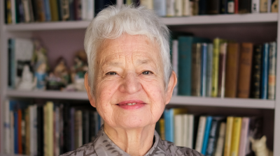 An author stood in front of a book case wearing a grey patterned shirt