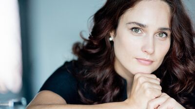A woman with long dark curly hair resting her chin on her finger, she has green eyes and pale skin
