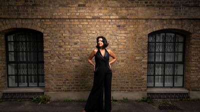 A woman wearing a black jumpsuit, stood against a brick wall