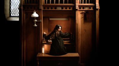 A person sat beneath a large organ with cream pipes, they are playing and turning to look at the camera behind