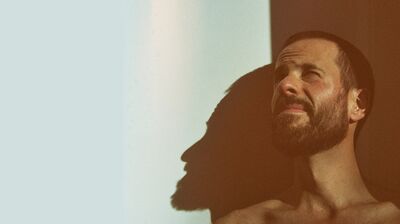 A person with close shaven hair and facial hair squints up sunshine with their shadow falling on the wall behind