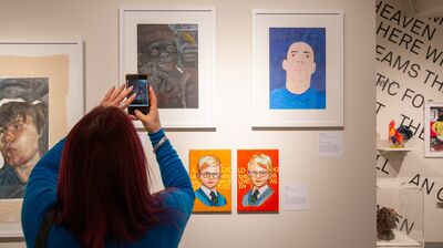 A person with long red hair wearing a blue sweater holds up her phone to photograph a picture within the Koestler Arts exhibition