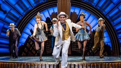 A theatrical performance with a male dancer in a white suit and hat leading a line of five dancers in 1920s-style costumes against a blue Art Deco backdrop.