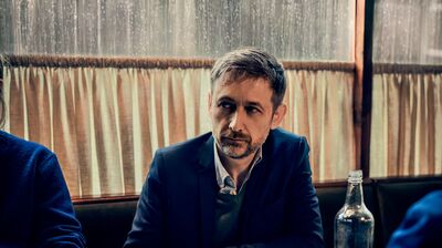 A man in a dark blue blazer and green sweater vest sits at a café table with a cup of coffee, looking contemplative.