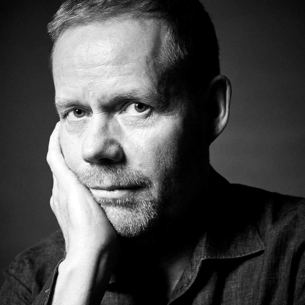 A black and white portrait of a man resting his head on his hand, looking directly at the camera with a serious expression.
