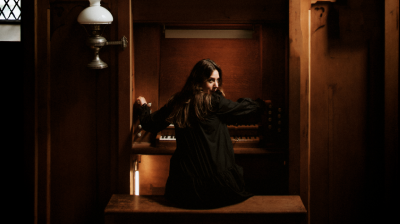 A person sat beneath a large organ with cream pipes, they are playing and turning to look at the camera behind
