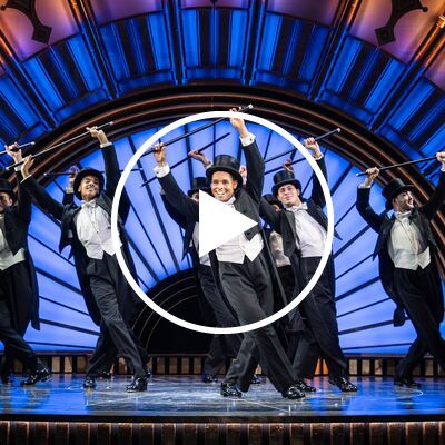 A musical theater ensemble of seven men in tuxedos and top hats dance with canes against a blue and gold arched backdrop.