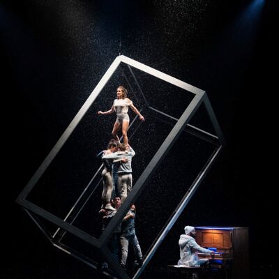 A dramatic acrobatic performance on a dark stage with simulated rain. A woman stands atop a human pyramid inside a large, tilted white cube frame, while a man plays a piano.