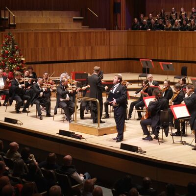 Orchestra performing on large wooden stage with a choir behind on a higher platform at the Royal Festival Hall