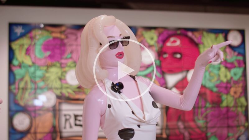 Performance artist Pademonia – a living sculpture of an exaggerated cartoon-like woman with blond hair, sunglasses and bright red lips, points towards a Gilbert & George picture within the Hayward Gallery